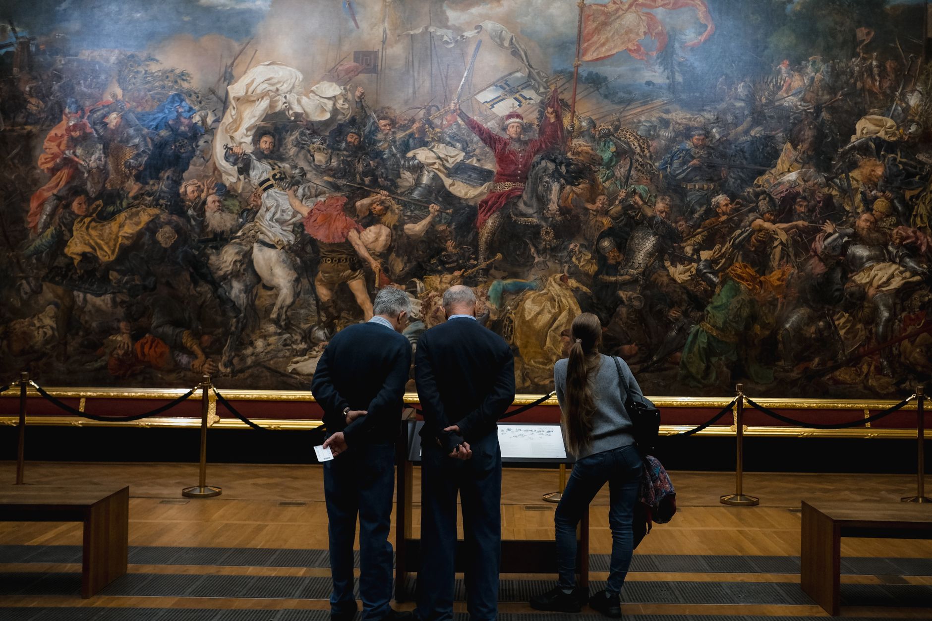 People viewing a large, detailed historical battle painting in a museum setting.