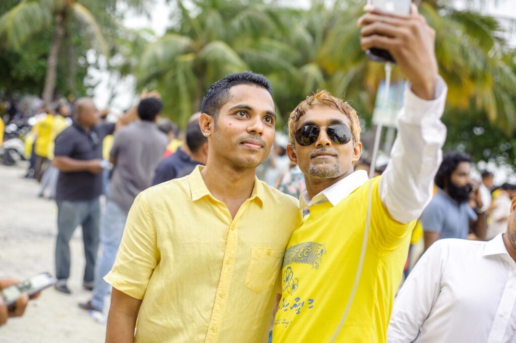 Two men taking a selfie during an outdoor gathering with a crowd in the background under palm trees.
