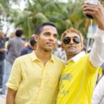 Two men taking a selfie during an outdoor gathering with a crowd in the background under palm trees.