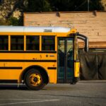 A classic yellow school bus parked on a city street with a rustic wooden building behind it.