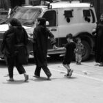 Black and white image capturing people walking past a police vehicle in an urban setting.