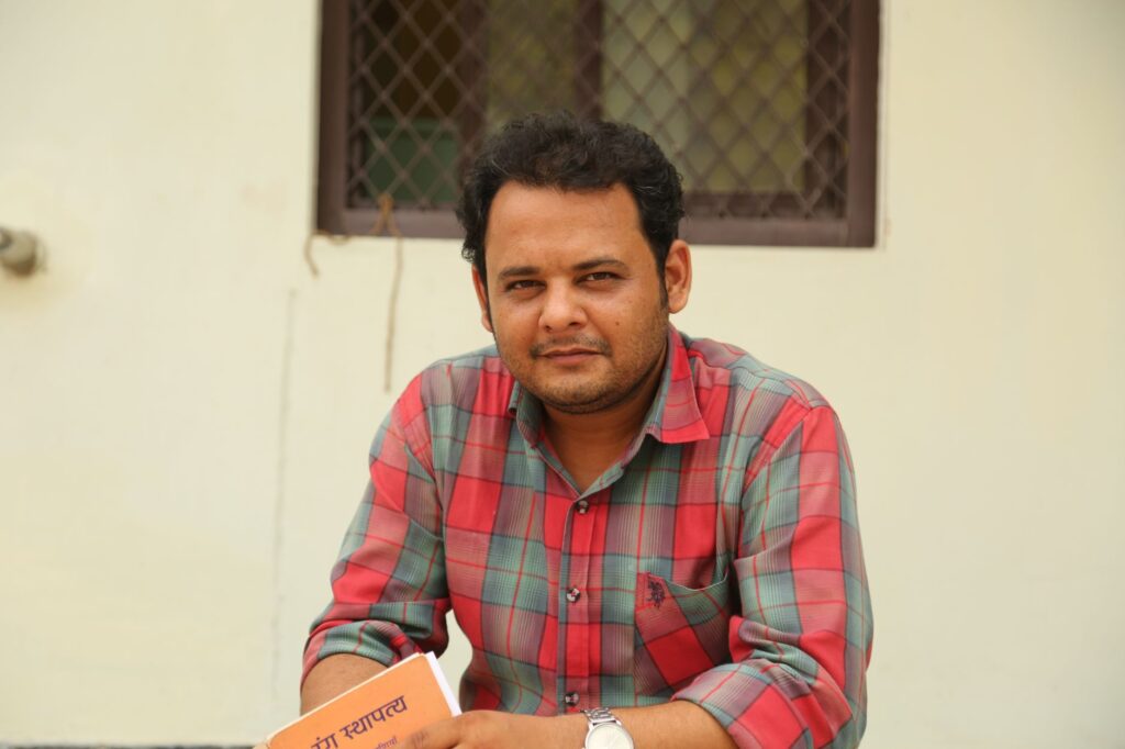 Casually dressed man with a book poses outdoors near a window wall.