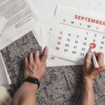 Hands holding a calendar with September dates encircled, surrounded by documents on a carpet.