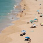 Aerial view of a crowded sandy beach with sunbathers and colorful umbrellas on a sunny day.