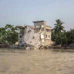 A damaged building collapsing by a tropical riverbank with palm trees and vegetation.