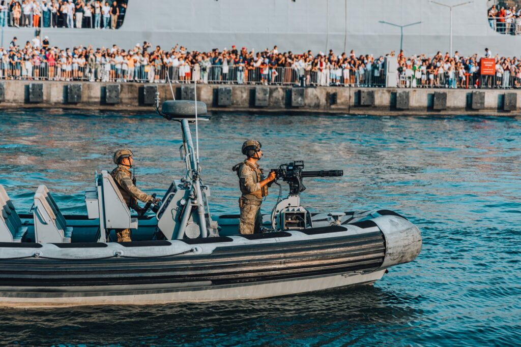 A military boat with armed soldiers patrolling near a crowded pier during a public event.