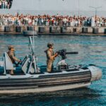 A military boat with armed soldiers patrolling near a crowded pier during a public event.