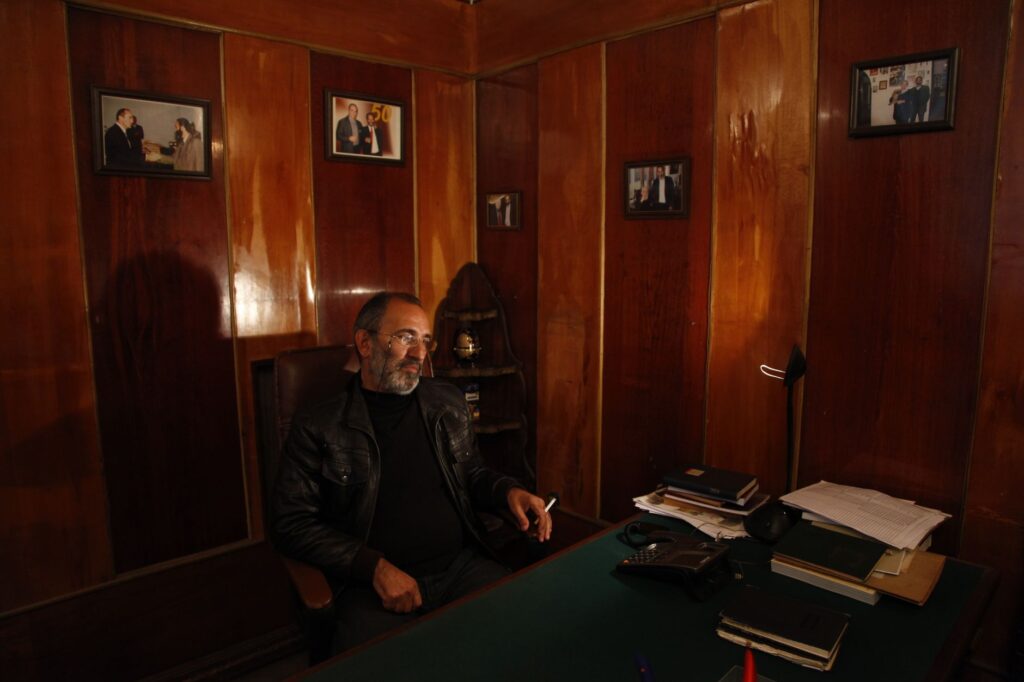 A mature man sits in a relaxed pose in a dimly lit, wood-paneled office, exuding an air of quiet authority.