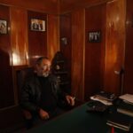 A mature man sits in a relaxed pose in a dimly lit, wood-paneled office, exuding an air of quiet authority.