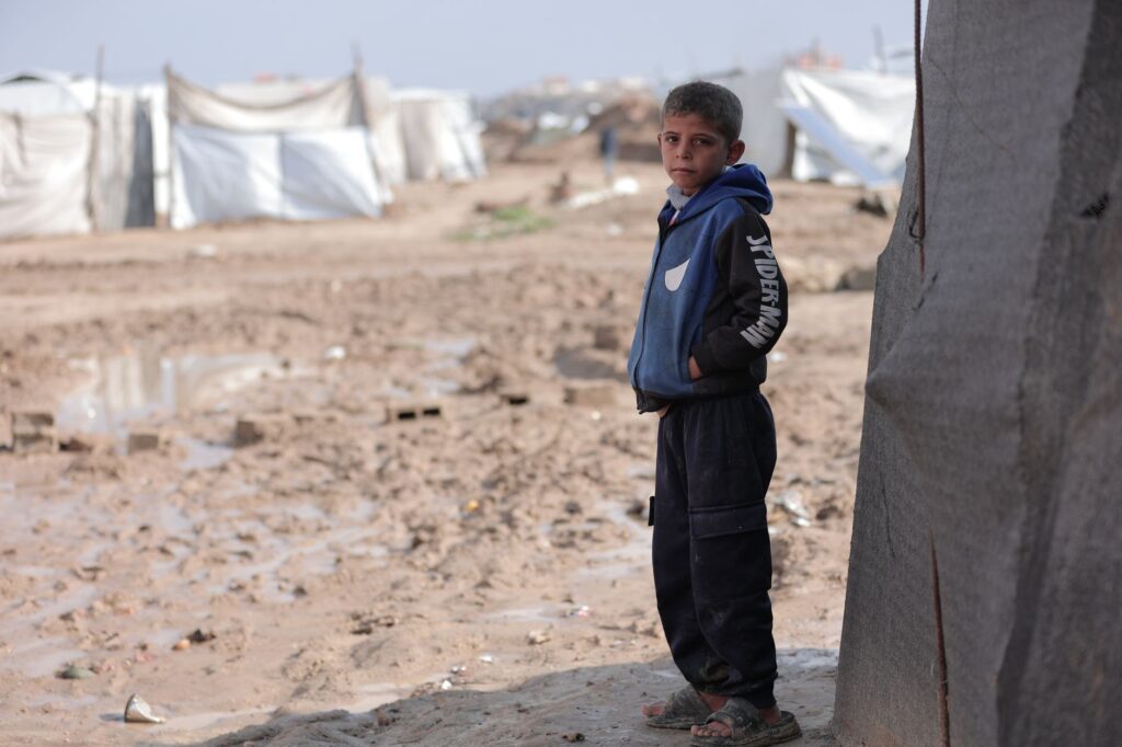 A boy stands near tents in a refugee camp on a cloudy day, looking thoughtful.