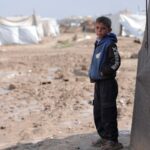 A boy stands near tents in a refugee camp on a cloudy day, looking thoughtful.