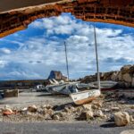 Sailboats rest on a pebbled shore beneath an arch at Calpe Harbor, with scenic cliffs in the background.