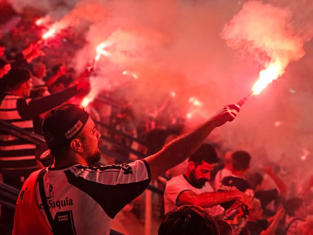 Energetic crowd celebrating with red flares at a sports event, creating a vibrant and passionate atmosphere.