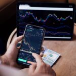 Close-up of a person trading stocks using a smartphone and a tablet.