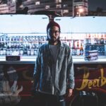 Young Asian male photographer in beanie and glasses wearing casual outfit standing near open street beer bar in night city