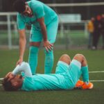 A soccer player lying on the grass field being consoled by a teammate in sportswear.