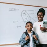 Smiling female teacher holding textbooks and standing near African American boy with backpack while looking at camera