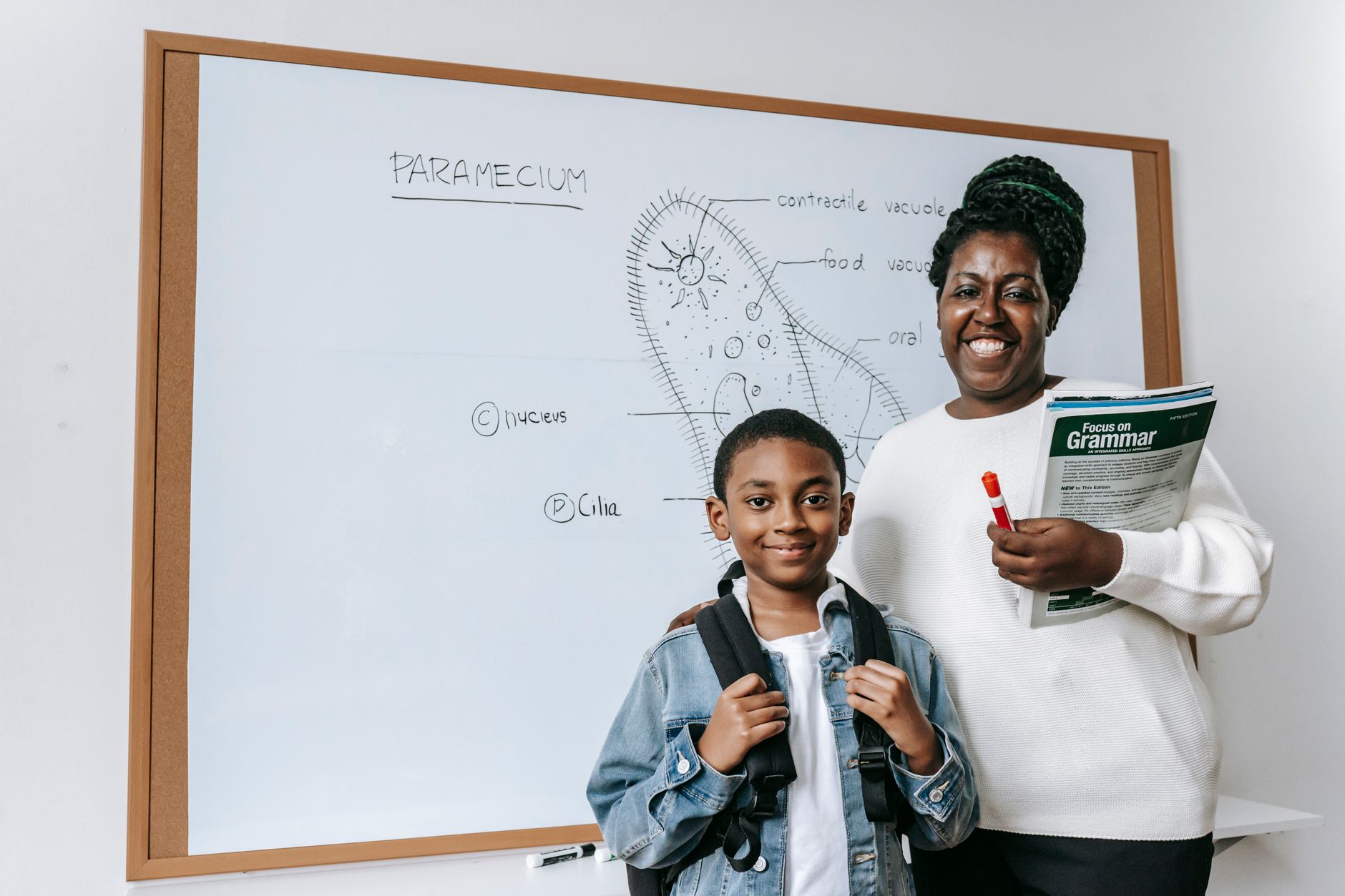 Smiling female teacher holding textbooks and standing near African American boy with backpack while looking at camera