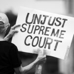 Black and white photo of a protester holding a sign saying 'Unjust Supreme Court' during a demonstration.