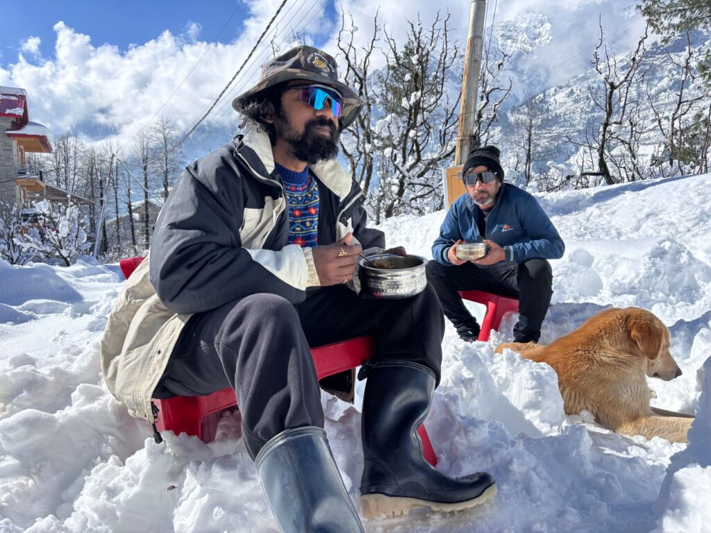 Two men seated in snowy landscape of Manali with a dog, enjoying a meal outdoors amidst a winter wonderland.