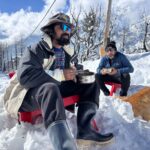 Two men seated in snowy landscape of Manali with a dog, enjoying a meal outdoors amidst a winter wonderland.