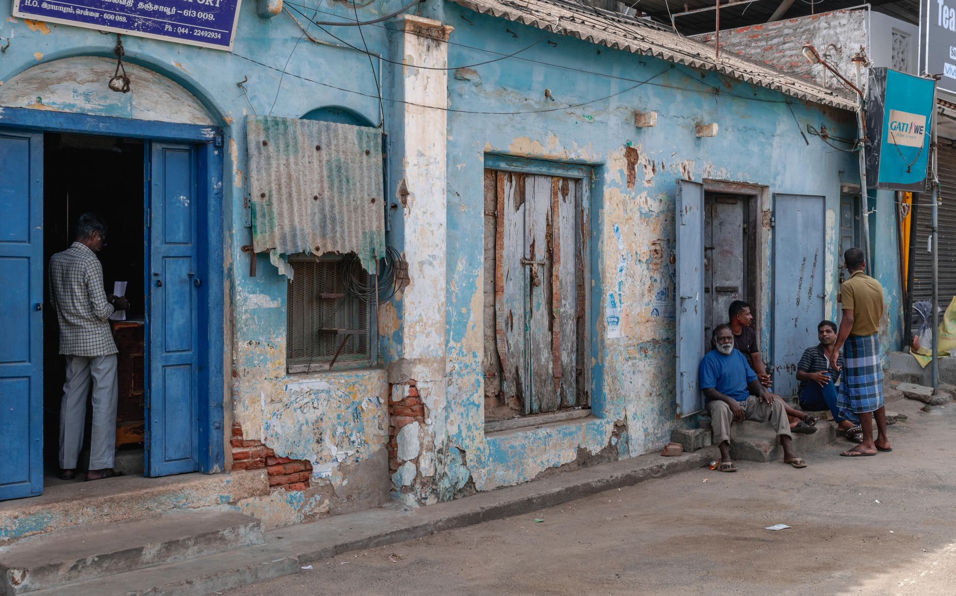 Local residents gather outside rustic buildings in Thanjavur, India, capturing daily life.