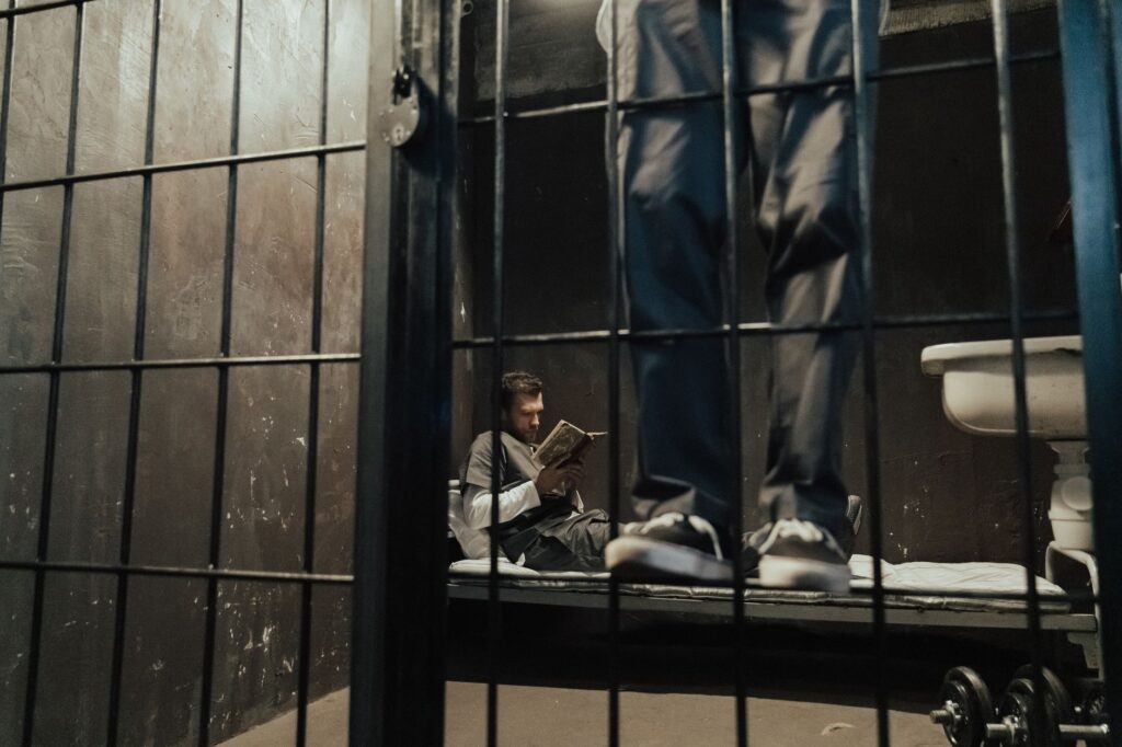 Inmate reading a book inside a prison cell while another stands nearby.