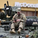 A soldier in camouflage resting on an armored vehicle, using a smartphone outside a McDonald's.