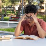 A young student studying at a table in an outdoor university campus setting, concentrating on his books.
