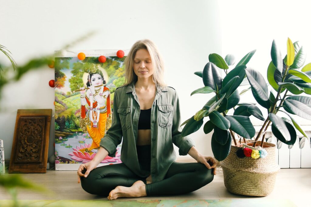 Woman meditating indoors with closed eyes, surrounded by vibrant plants and spiritual art for wellness.