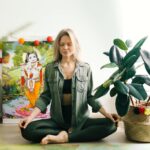 Woman meditating indoors with closed eyes, surrounded by vibrant plants and spiritual art for wellness.