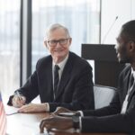 Two business professionals discussing and signing documents at a meeting with an American flag on the table.