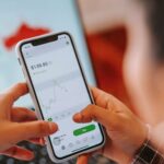 Crop anonymous trader using app on cellphone with graph and dollar price on screen in house