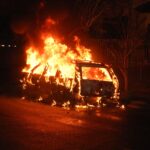 A vehicle is completely engulfed in flames, casting a dramatic glow on a dark street.