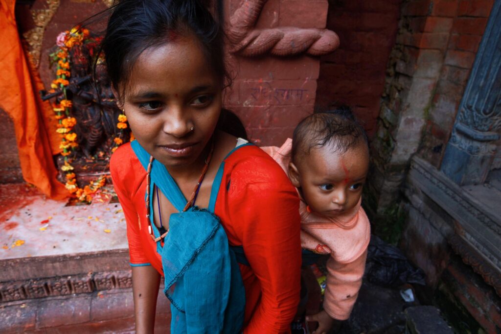 A South Asian woman carrying her baby in traditional attire at an ornate temple setting.