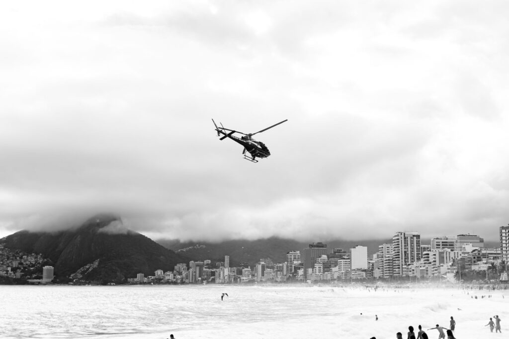 Black and white image of a helicopter flying over Rio de Janeiro's coastline with a mountainous backdrop.