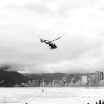 Black and white image of a helicopter flying over Rio de Janeiro's coastline with a mountainous backdrop.