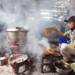 Street food vendor grills satay on skewers over open flames in a traditional market in Yogyakarta.