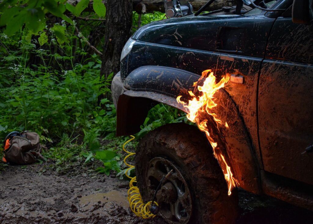 A close-up of a mud-splattered off-road vehicle on fire in a dense forest, creating a dramatic scene.