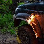 A close-up of a mud-splattered off-road vehicle on fire in a dense forest, creating a dramatic scene.