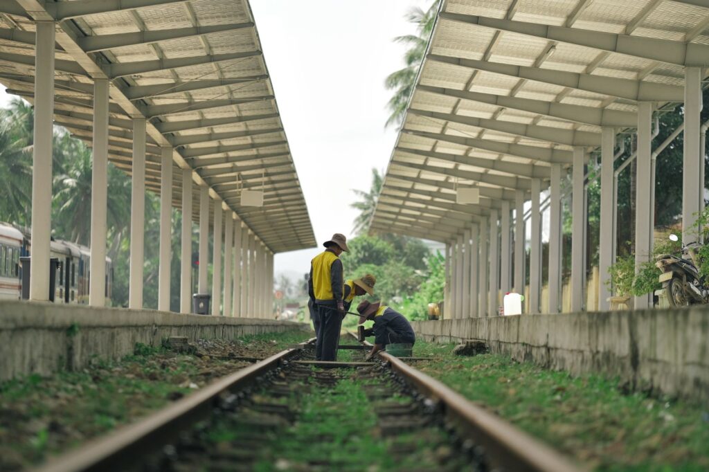Workers in hats maintain train tracks at a rural railway station. Sunny day setting.