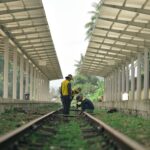 Workers in hats maintain train tracks at a rural railway station. Sunny day setting.