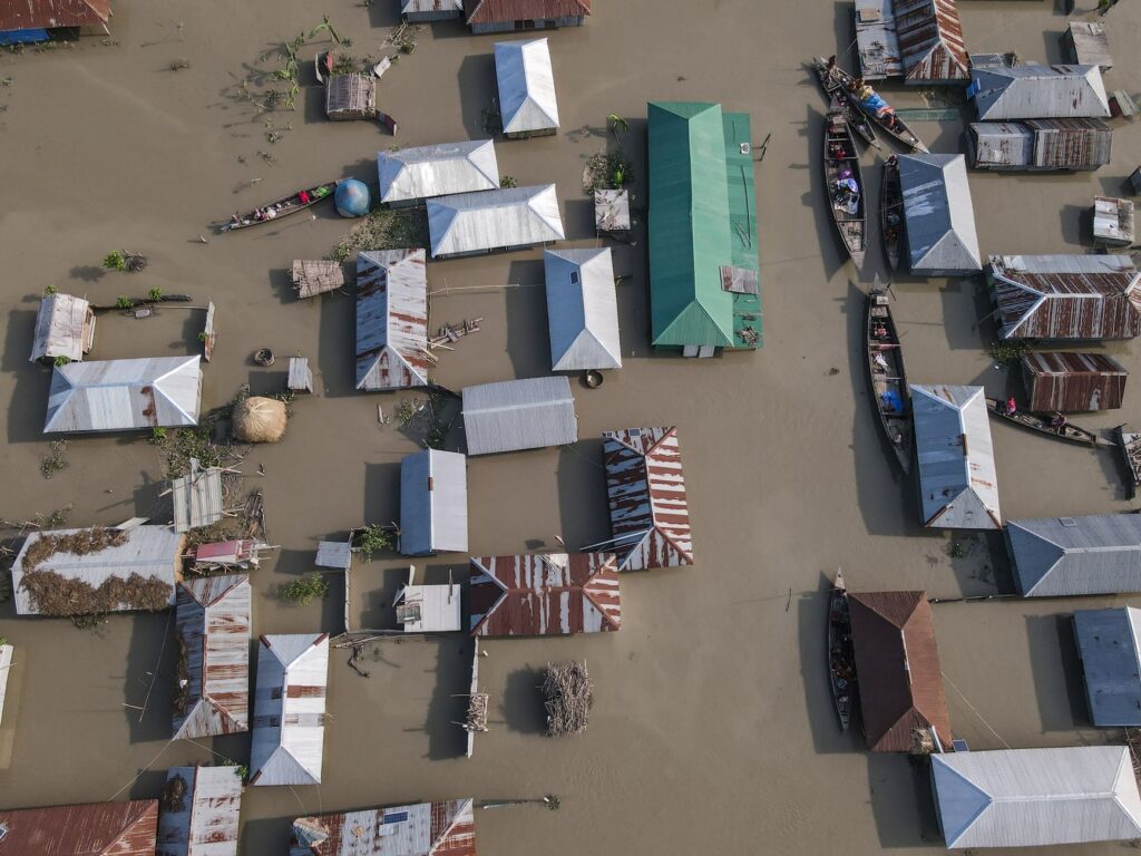 Aerial perspective of a village submerged in floodwaters, highlighting tin-roofed houses and boats.
