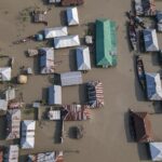 Aerial perspective of a village submerged in floodwaters, highlighting tin-roofed houses and boats.