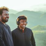 Two men smiling and enjoying a scenic mountain view outdoors, wearing jackets and headphones.