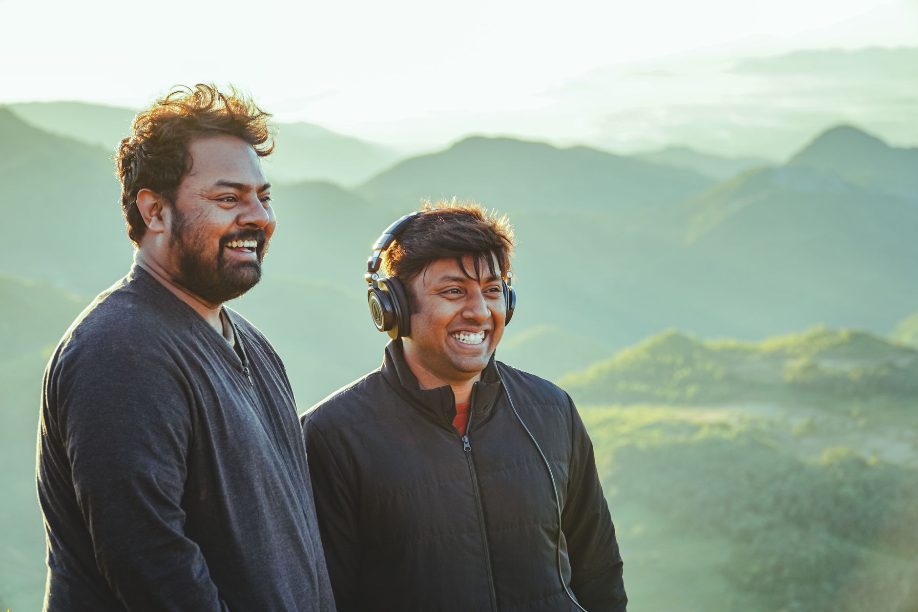 Two men smiling and enjoying a scenic mountain view outdoors, wearing jackets and headphones.
