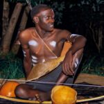 A young African man sitting outdoors with traditional tribal body paint and accessories at night in Uganda.
