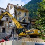 Excavator demolishing building against a scenic mountain backdrop, showcasing urban construction.