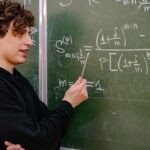 A young male educator explaining complex math equations at a university classroom blackboard.