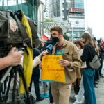 Young man holds protest sign during street demonstration against war, covered by media.